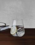 NUDE Salute tumbler with water, ice and lemon slice beside a book and reading glasses on a wooden table
