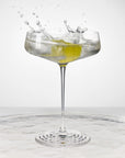NUDE | Tom Dixon Splash Coupe glass with a clear cocktail and olive garnish splashing, set against a white background on a reflective surface.