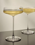 Two NUDE | Tom Dixon Splash Coupe glasses filled with a pale gold sparkling drink, side by side on a dark reflective surface.