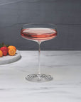 NUDE | Tom Dixon Splash Coupe glass filled with a pale pink drink on a marble surface, with strawberries, blueberries, and citrus fruit on a stone board blurred in the background.