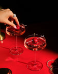 Two NUDE | Tom Dixon Splash coupe glasses filled with pink cocktails and cherry garnishes on a red reflective surface, with a hand placing a cherry into one glass and gold jewelry in the background