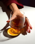 Overhead close-up of a hand holding the NUDE | Tom Dixon Splash Lowball glass filled with amber liquid, warm light casting long shadows across a white surface.