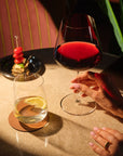 A hand holds a Sera Burgundy glass of red wine beside a Sera Tumbler of water and lemon, with a skewered appetizer on a small plate nearby.
