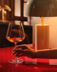A hand holds a Sera Burgundy glass of orange wine on a glossy red table, lit by a warm table lamp in a dark wood bar setting.