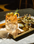 A styled cocktail in a Sera tumbler glass with ice, garnished with green olives, a lemon twist, and a rosemary sprig, served on a gold tray alongside a small glass Silhouette bowl of green olives on a stone tabletop.
