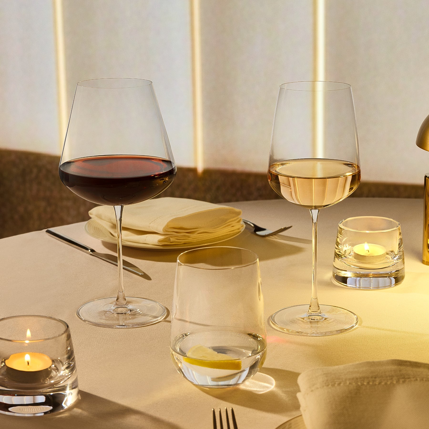 Two NUDE Sera wine glasses on a softly lit dining table, one filled with red wine and one with white wine, alongside candle holders, folded napkins, and cutlery.