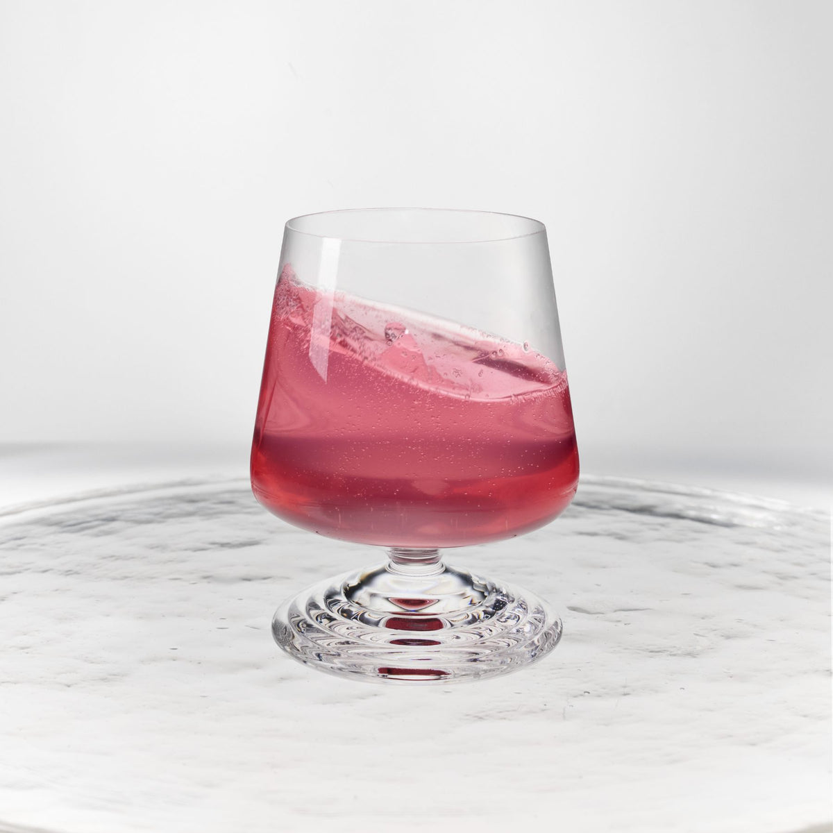 NUDE | Tom Dixon Splash Lowball glass with pink liquid actively splashing, set on a reflective marble surface.