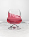 NUDE | Tom Dixon Splash Lowball glass with pink liquid actively splashing, set on a reflective marble surface.