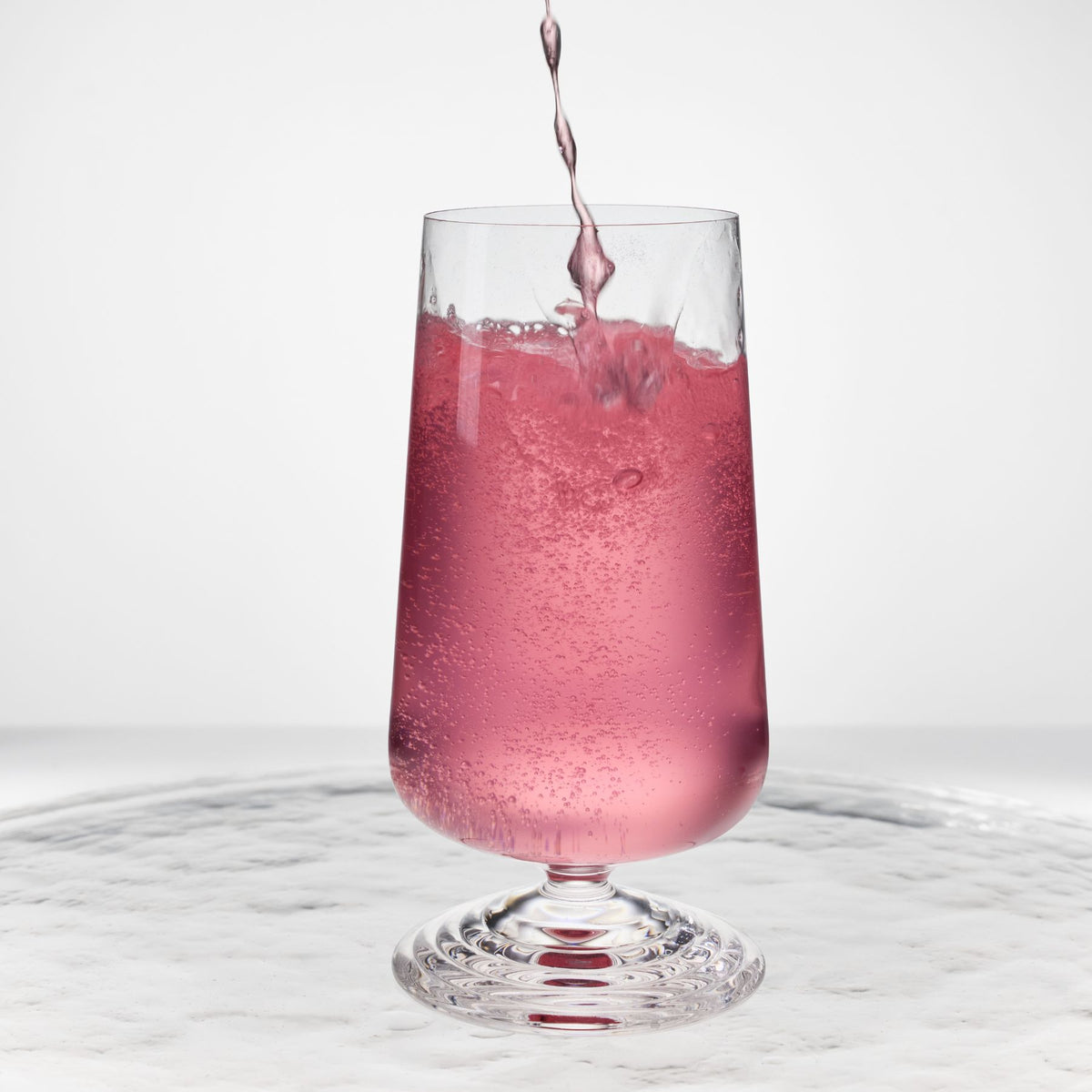 NUDE | Tom Dixon Splash Highball glass with pink sparkling liquid actively pouring in, set on a reflective marble surface.