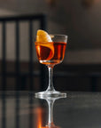 A NUDE BarGiani welcome drink glass filled with an amber-orange cocktail and garnished with a curled orange peel, set on a dark reflective surface with a blurred indoor background.