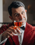 Giorgio BarGiani in a red velvet blazer, white shirt, and black bow tie holds up the NUDE Welcome drink glass with amber liquid, likely a whiskey or liqueur, in a dimly lit indoor setting.