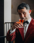 Giorgio Bargiani in a red velvet tuxedo jacket, white shirt, and black bow tie holds up his NUDE welcome drink glass with amber liquid, looking directly at the camera in an indoor setting.