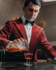 Giorgio BarGiani in a red velvet blazer, white dress shirt, and black bow tie pours an amber-colored cocktail from his NUDE doublepour mixing glass shaker into the new NUDE welcome drink glass on a dark marble table.