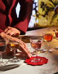 A close-up lifestyle shot of a person in a burgundy blazer placing a pink cocktail on a scalloped red coaster on a white marble bar counter. Four other NUDE Bargiani welcome drink glasses are lined up behind it on a warm, dimly lit bar table.