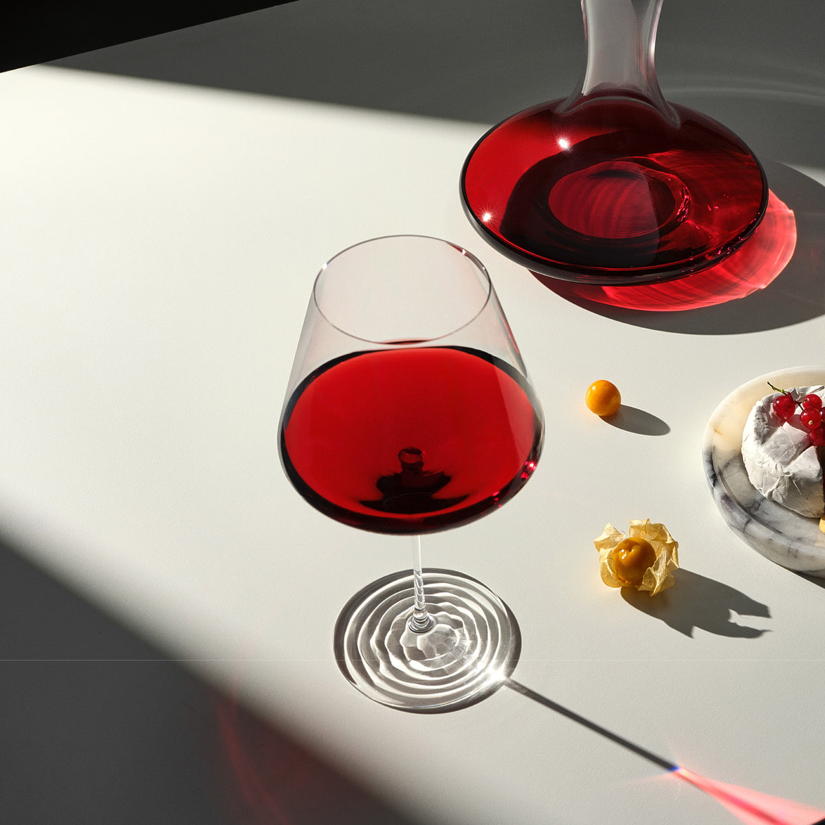NUDE Splash red wine glass with the ripple foot base, filled with red wine, positioned on a sunlit white surface next to a NUDE Balance wine carafe holding red wine. Physalis fruit, a redcurrant sprig, and a wheel of cheese sit nearby, with strong diagonal shadow and light across the table.