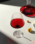 NUDE Splash red wine glass with the ripple foot base, filled with red wine, positioned on a sunlit white surface next to a NUDE Balance wine carafe holding red wine. Physalis fruit, a redcurrant sprig, and a wheel of cheese sit nearby, with strong diagonal shadow and light across the table.