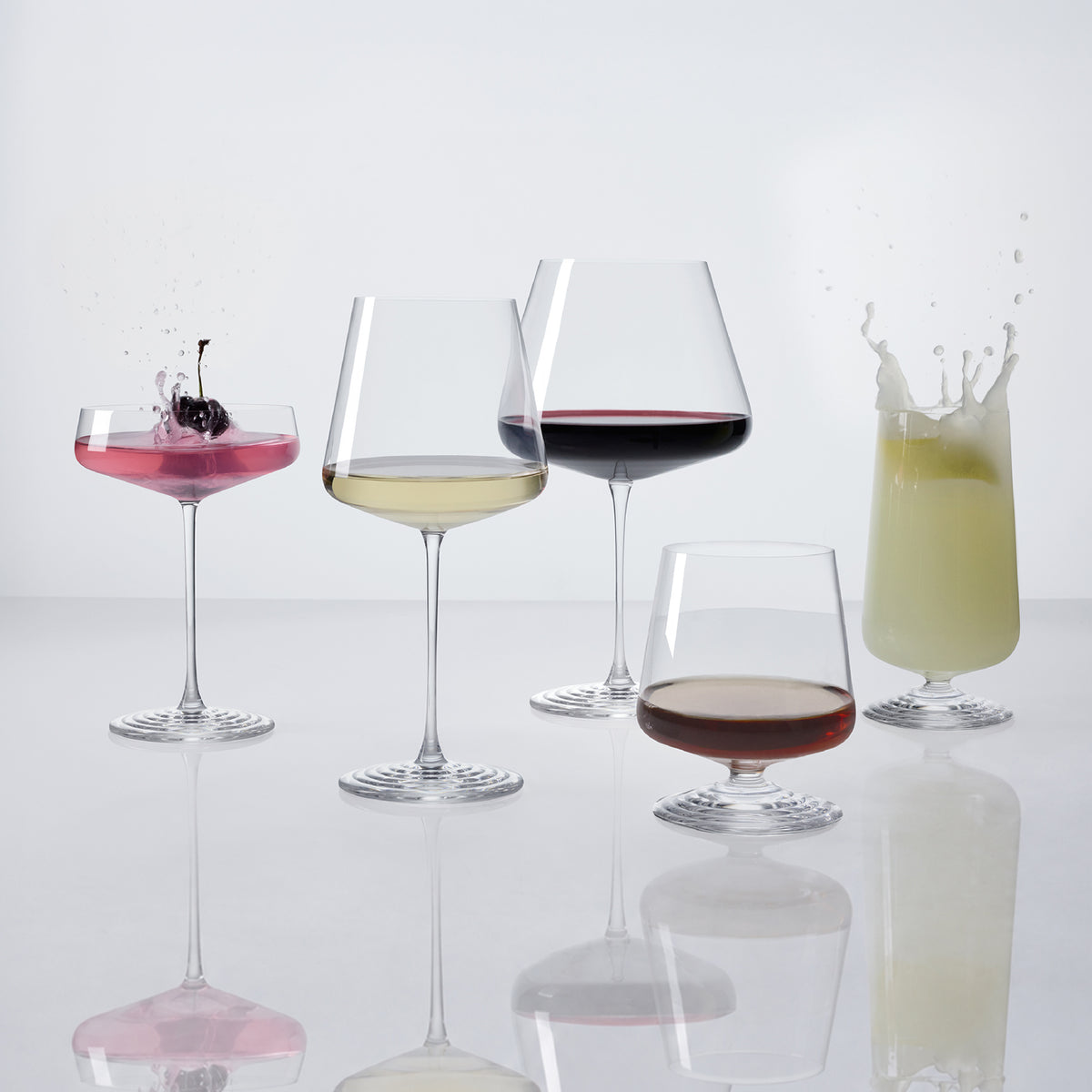 The five NUDE | Tom Dixon Splash glasses in a row, each filled: coupe with pink cocktail and cherry garnish splashing, white wine, red wine, lowball with amber spirit, and highball with a pale green drink splashing, all on a white reflective surface.