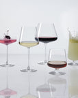 The five NUDE | Tom Dixon Splash glasses in a row, each filled: coupe with pink cocktail and cherry garnish splashing, white wine, red wine, lowball with amber spirit, and highball with a pale green drink splashing, all on a white reflective surface.