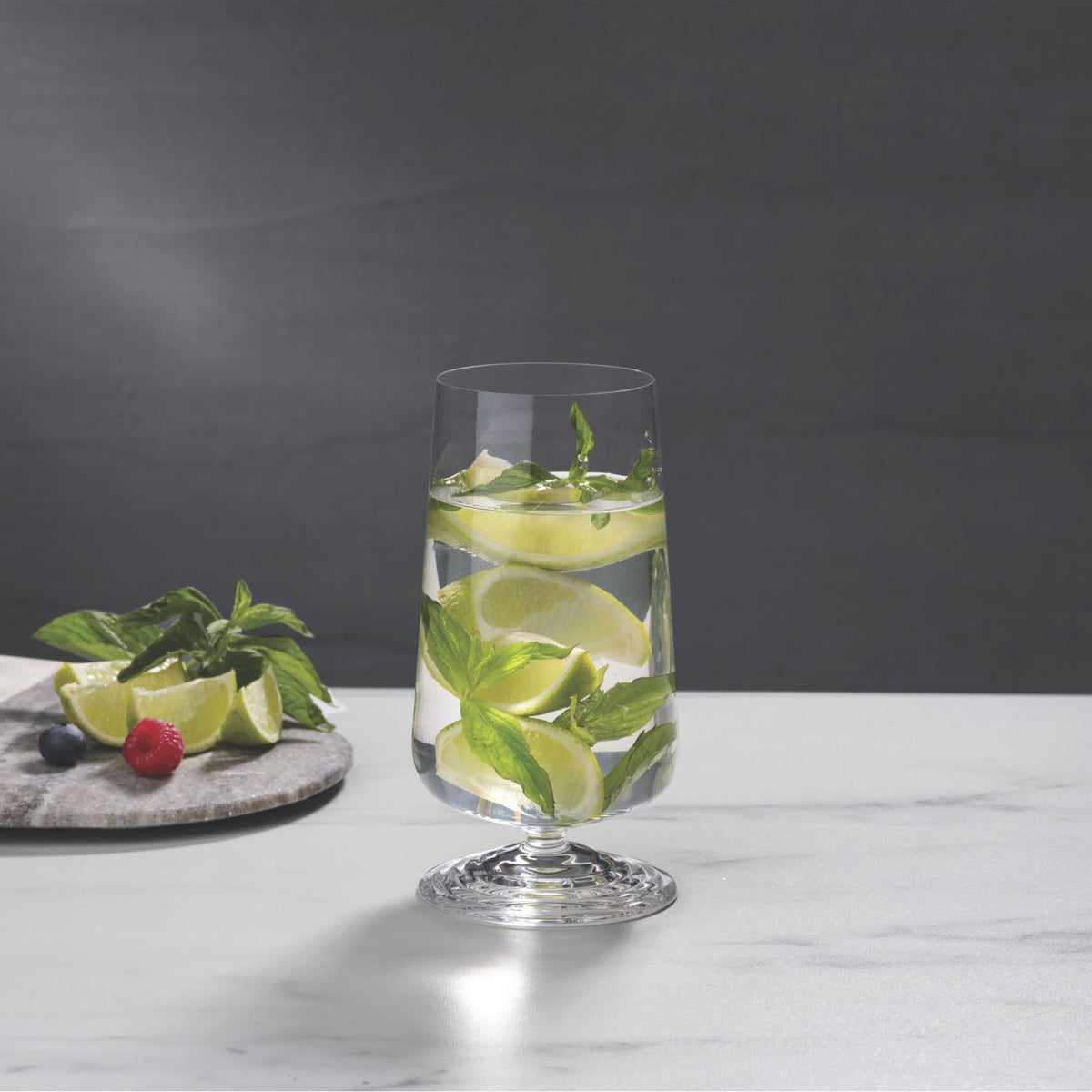 NUDE | Tom Dixon Splash Highball glass filled with water, lime wedges, and mint leaves on a marble surface, with more mint, lime, and berries on a stone board blurred in the background.