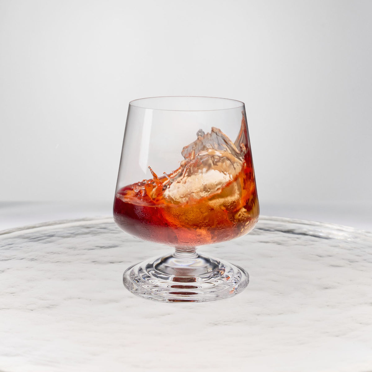 NUDE | Tom Dixon Splash Lowball glass with amber spirit and ice splashing, set on a reflective marble surface.