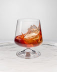 NUDE | Tom Dixon Splash Lowball glass with amber spirit and ice splashing, set on a reflective marble surface.