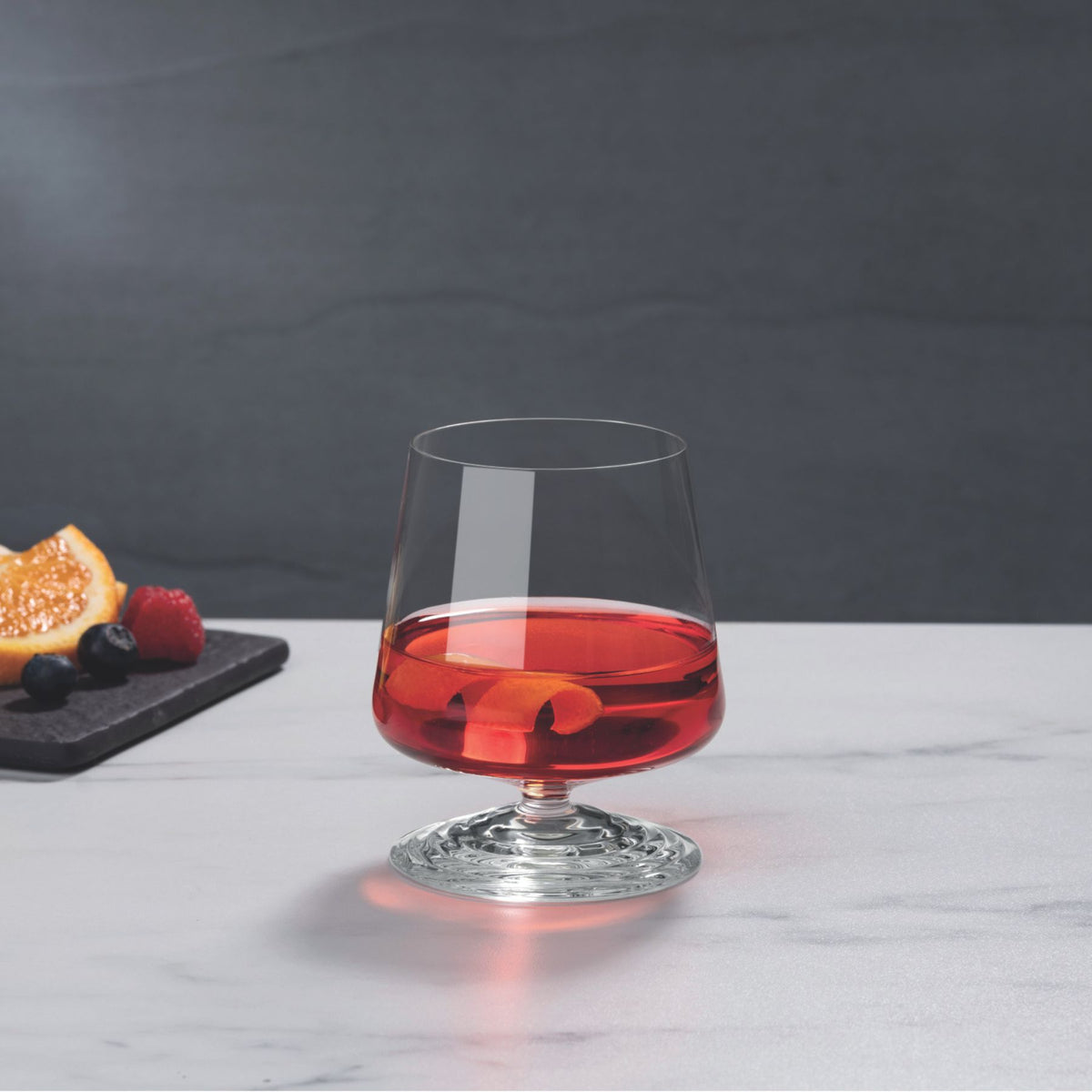 NUDE | Tom Dixon Splash Lowball glass filled with a red drink and citrus twist garnish on a marble surface, with orange slices and berries on a dark board blurred in the background.