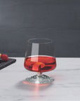 NUDE | Tom Dixon Splash Lowball glass filled with a red drink and citrus twist garnish on a marble surface, with orange slices and berries on a dark board blurred in the background.