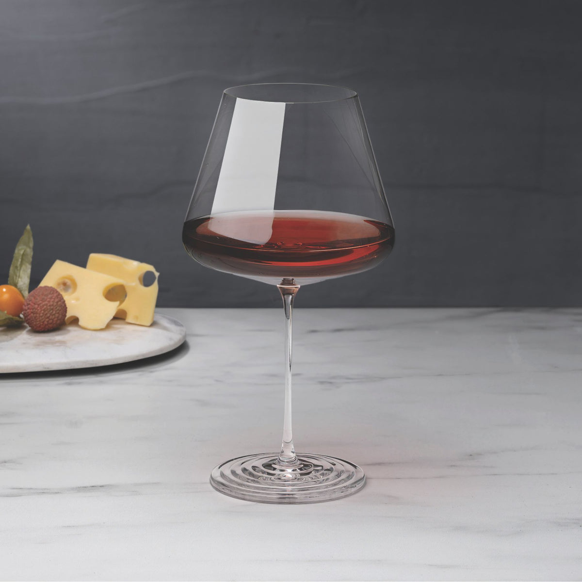 NUDE Splash Red Wine glass filled with red wine on a marble surface, with a cheese and fruit board blurred in the background against a dark backdrop.