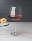 NUDE Splash Red Wine glass filled with red wine on a marble surface, with a cheese and fruit board blurred in the background against a dark backdrop.