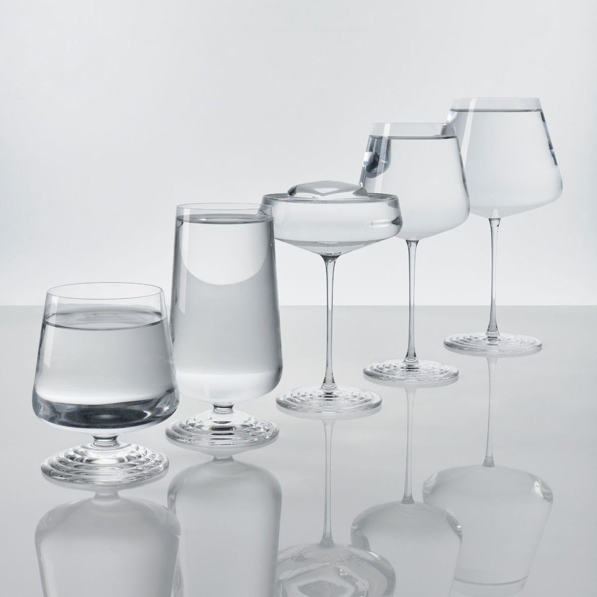 The five NUDE Splash glasses filled with water in an ascending row, including the Splash Red Wine glass, each showing the signature ripple foot base.