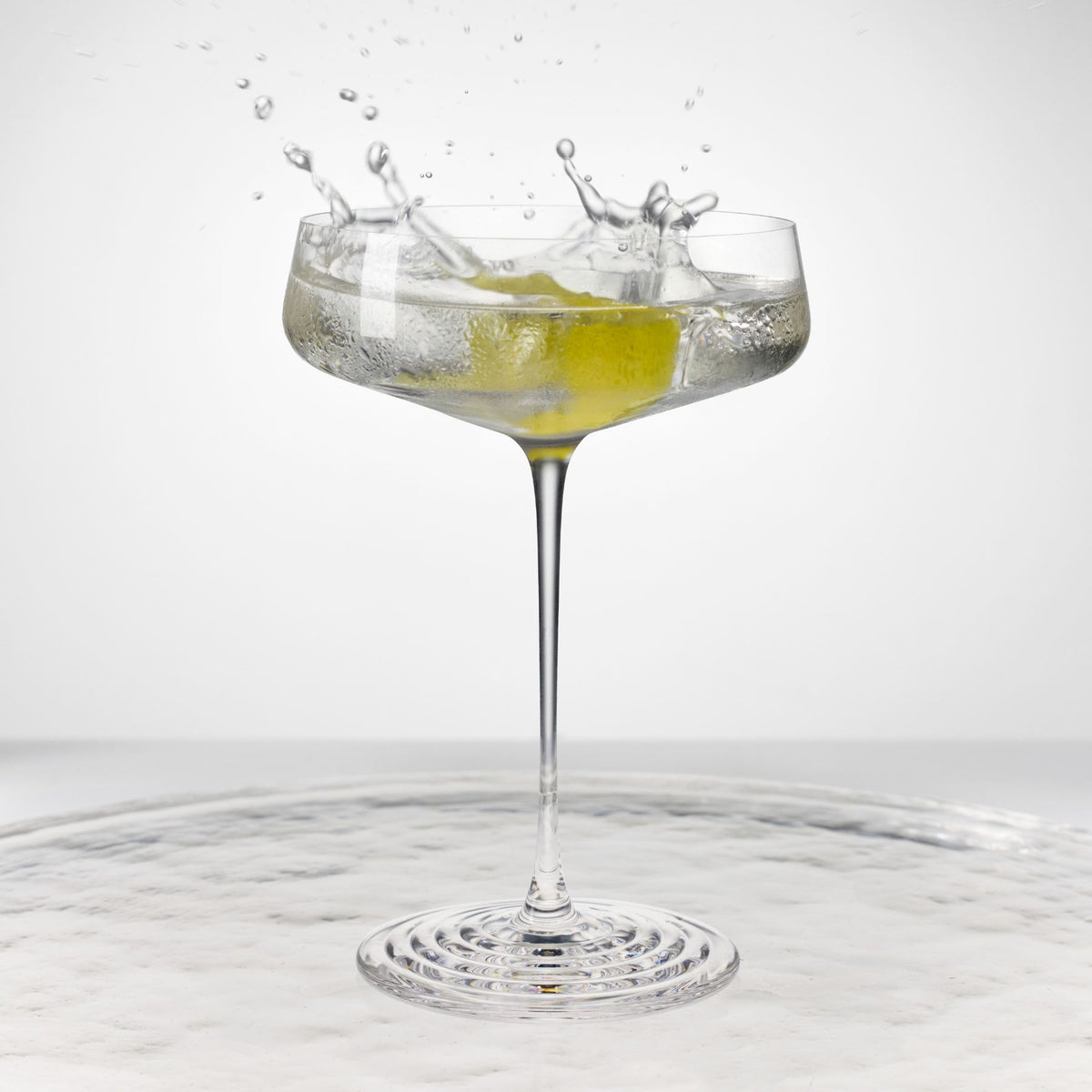 NUDE | Tom Dixon Splash Coupe glass with a clear cocktail and olive garnish splashing, set against a white background on a reflective surface.