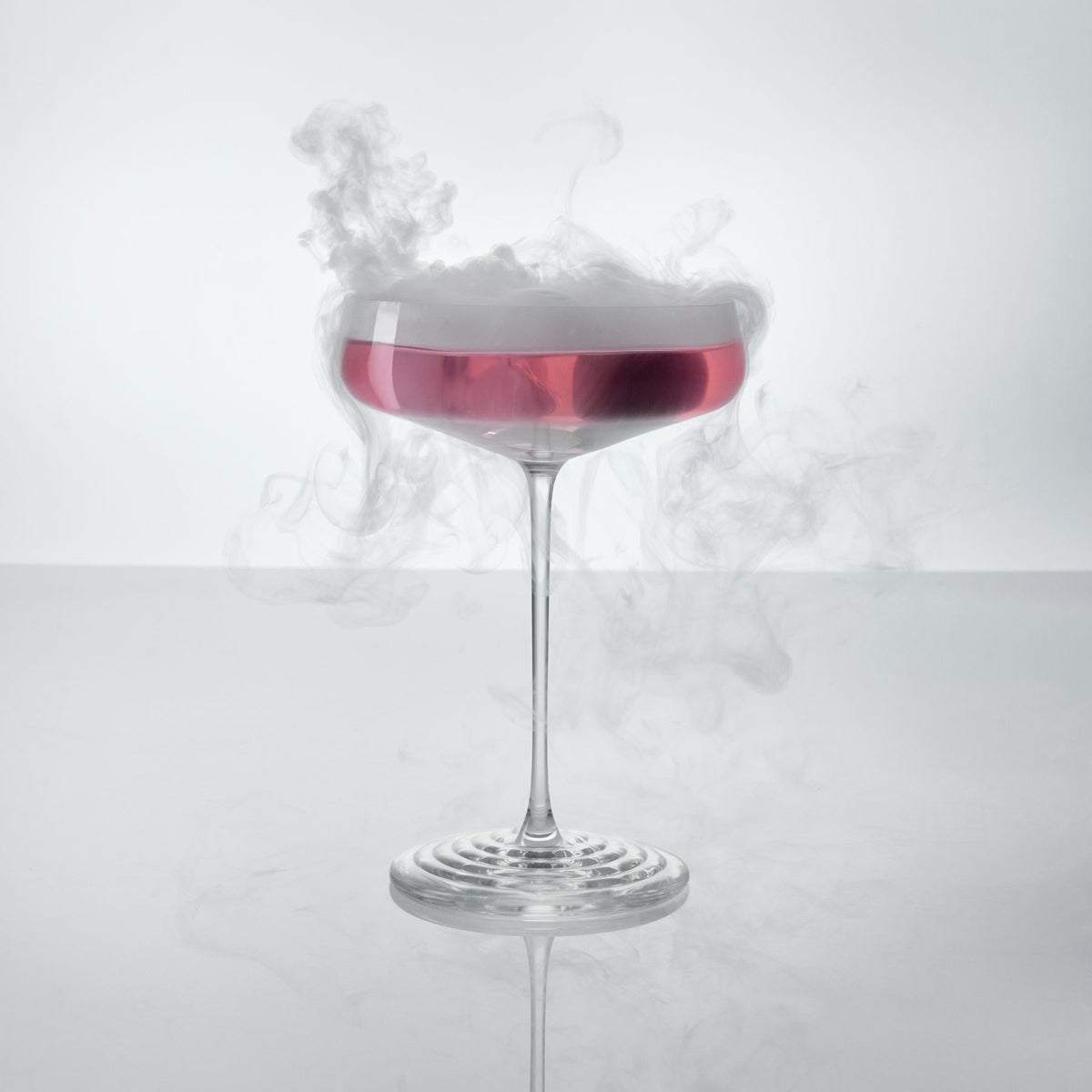 NUDE | Tom Dixon Splash Coupe glass filled with a pink drink, with smoke swirling above the rim against a white background.