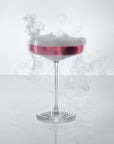 NUDE | Tom Dixon Splash Coupe glass filled with a pink drink, with smoke swirling above the rim against a white background.