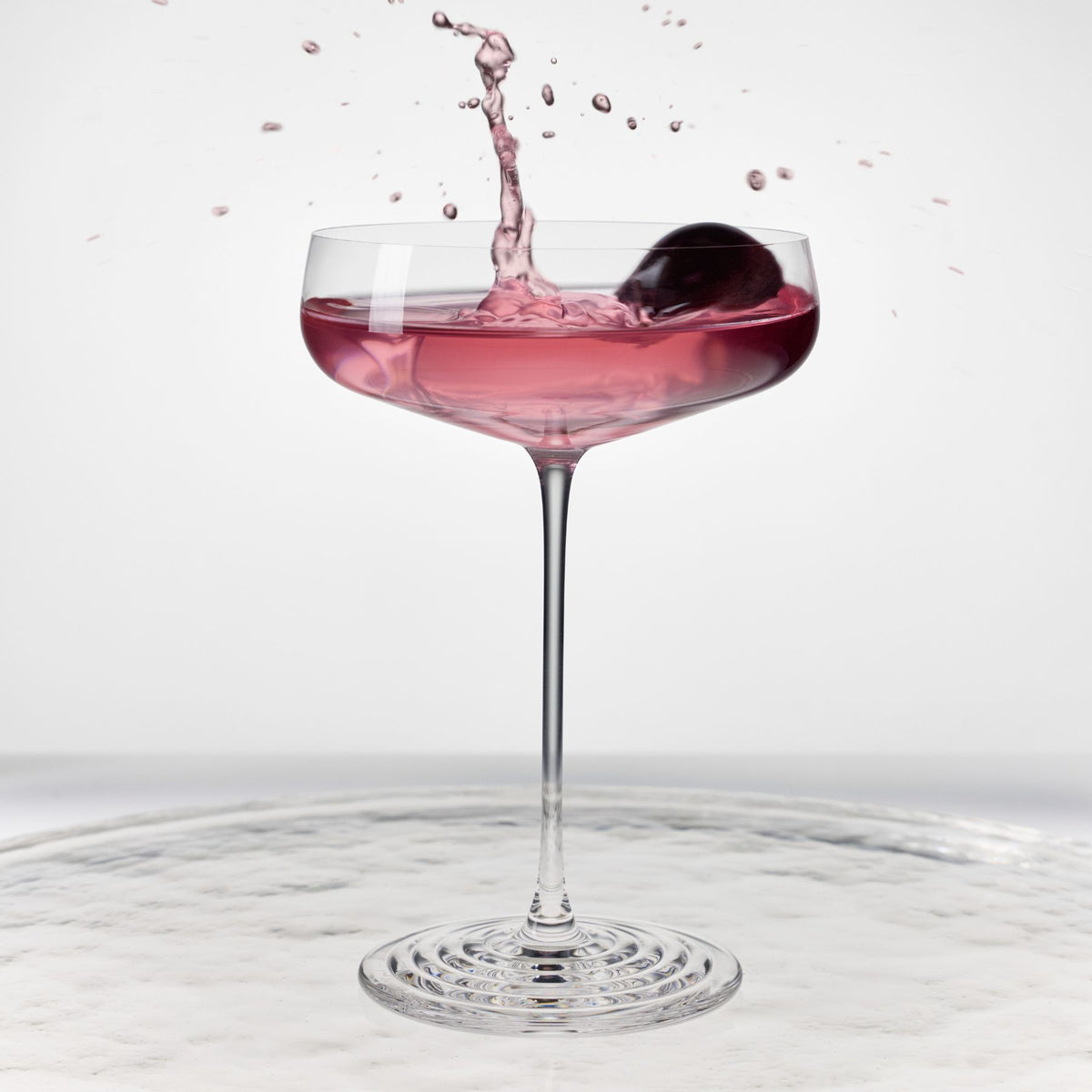 NUDE | Tom Dixon Splash coupe glass with pink cocktail and cherry garnish splashing, set against a white background.