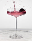 NUDE | Tom Dixon Splash coupe glass with pink cocktail and cherry garnish splashing, set against a white background.