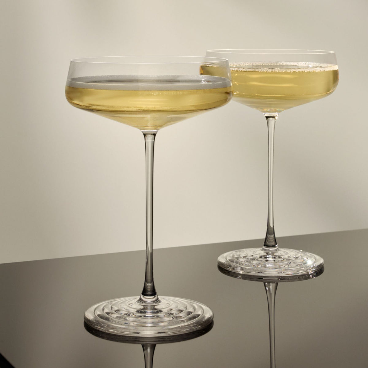 Two NUDE | Tom Dixon Splash Coupe glasses filled with a pale gold sparkling drink, side by side on a dark reflective surface.