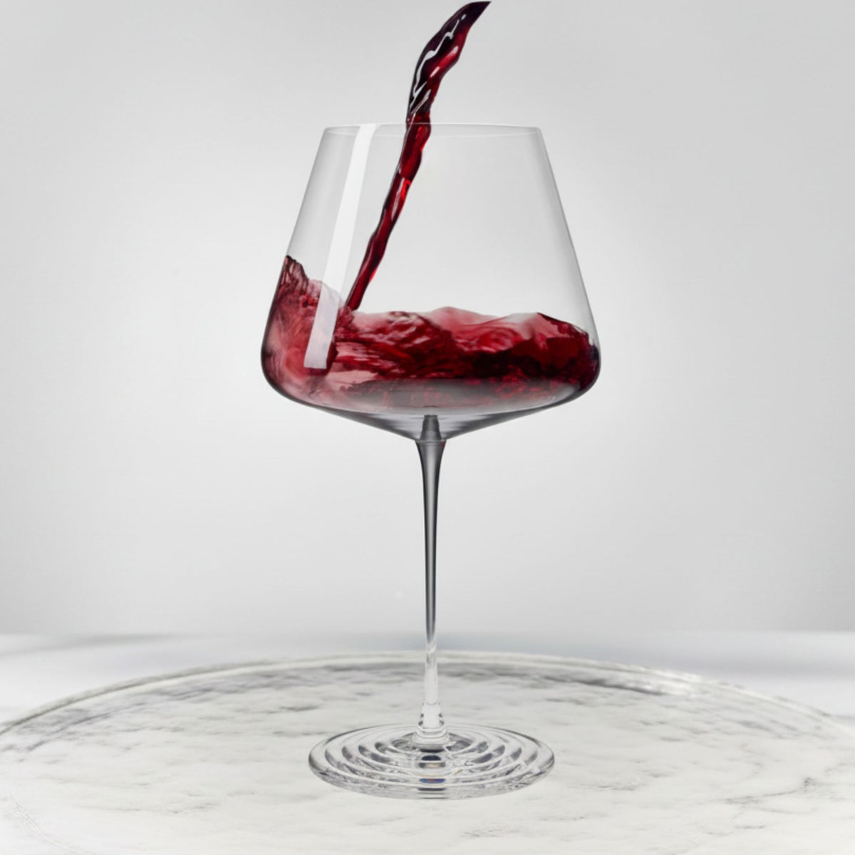 NUDE Splash Red Wine glass with red wine actively pouring in and splashing against the bowl, set on a reflective marble surface.