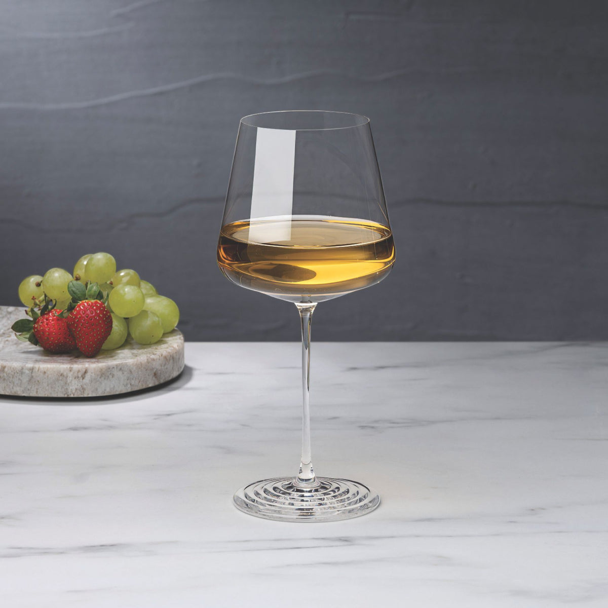 NUDE | Tom Dixon Splash White Wine glass filled with white wine on a marble surface, with grapes and strawberries on a stone board blurred in the background