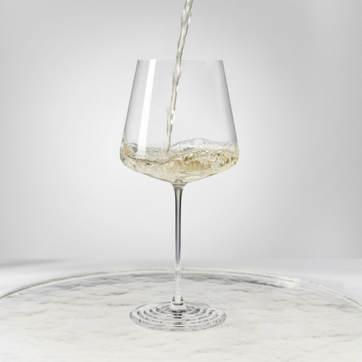 NUDE | Tom Dixon Splash White Wine glass with white wine actively pouring in and splashing against the bowl, set on a reflective marble surface.