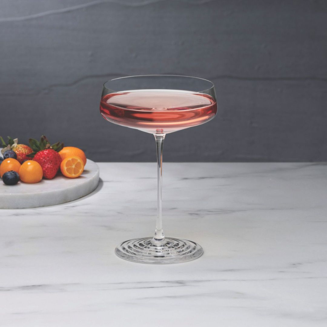 NUDE | Tom Dixon Splash Coupe glass filled with a pale pink drink on a marble surface, with strawberries, blueberries, and citrus fruit on a stone board blurred in the background.