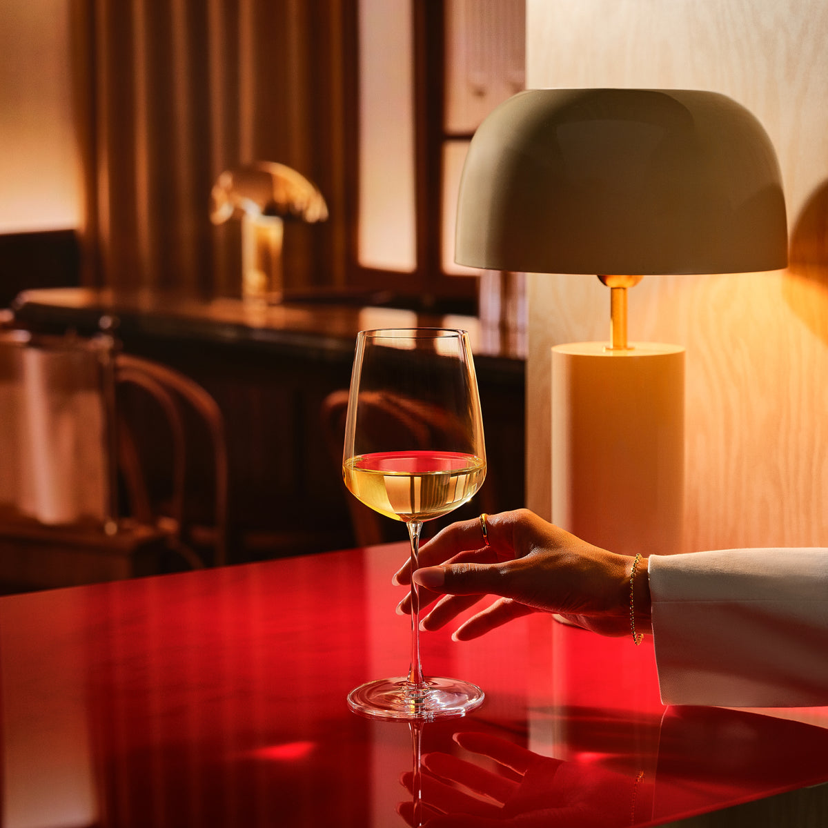 A hand in a white sleeve holds a NUDE Sera wine glass with a small amount of white wine on a glossy red table, with a warm lamp and dark interior softly blurred in the background.