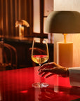 A hand in a white sleeve holds a NUDE Sera wine glass with a small amount of white wine on a glossy red table, with a warm lamp and dark interior softly blurred in the background.
