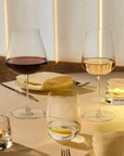 Two NUDE Sera wine glasses on a softly lit dining table, one filled with red wine and one with white wine, alongside candle holders, folded napkins, and cutlery.