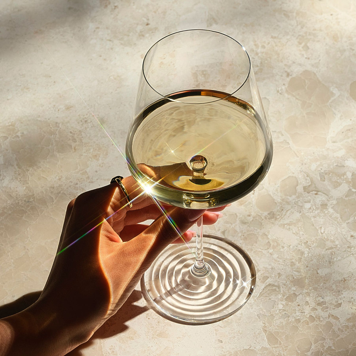 Overhead close-up of a hand holding the NUDE | Tom Dixon Splash White Wine glass filled with white wine, with sunlight creating a lens flare across the marble surface below.