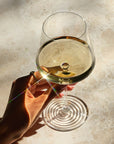 Overhead close-up of a hand holding the NUDE | Tom Dixon Splash White Wine glass filled with white wine, with sunlight creating a lens flare across the marble surface below.