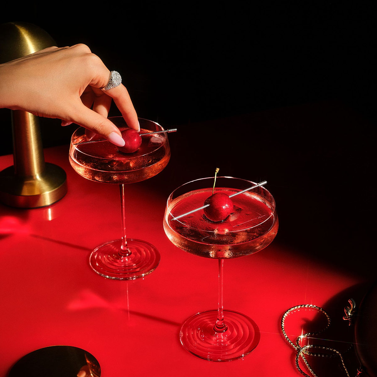 Two NUDE | Tom Dixon Splash coupe glasses filled with pink cocktails and cherry garnishes on a red reflective surface, with a hand placing a cherry into one glass and gold jewelry in the background