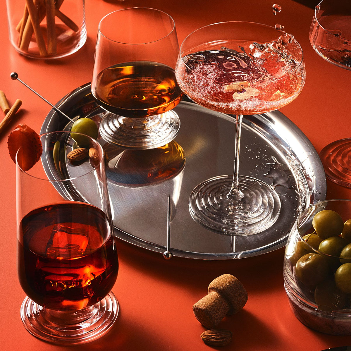 NUDE | Tom Dixon Splash glasses styled on a warm terracotta backdrop: a lowball with amber spirit and ice on a silver tray, a coupe with pink cocktail splashing and edible flower garnish, and a lowball with dark red drink in the foreground, surrounded by olives, cocktail picks, and cinnamon sticks.