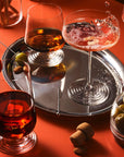 NUDE | Tom Dixon Splash glasses styled on a warm terracotta backdrop: a lowball with amber spirit and ice on a silver tray, a coupe with pink cocktail splashing and edible flower garnish, and a lowball with dark red drink in the foreground, surrounded by olives, cocktail picks, and cinnamon sticks.