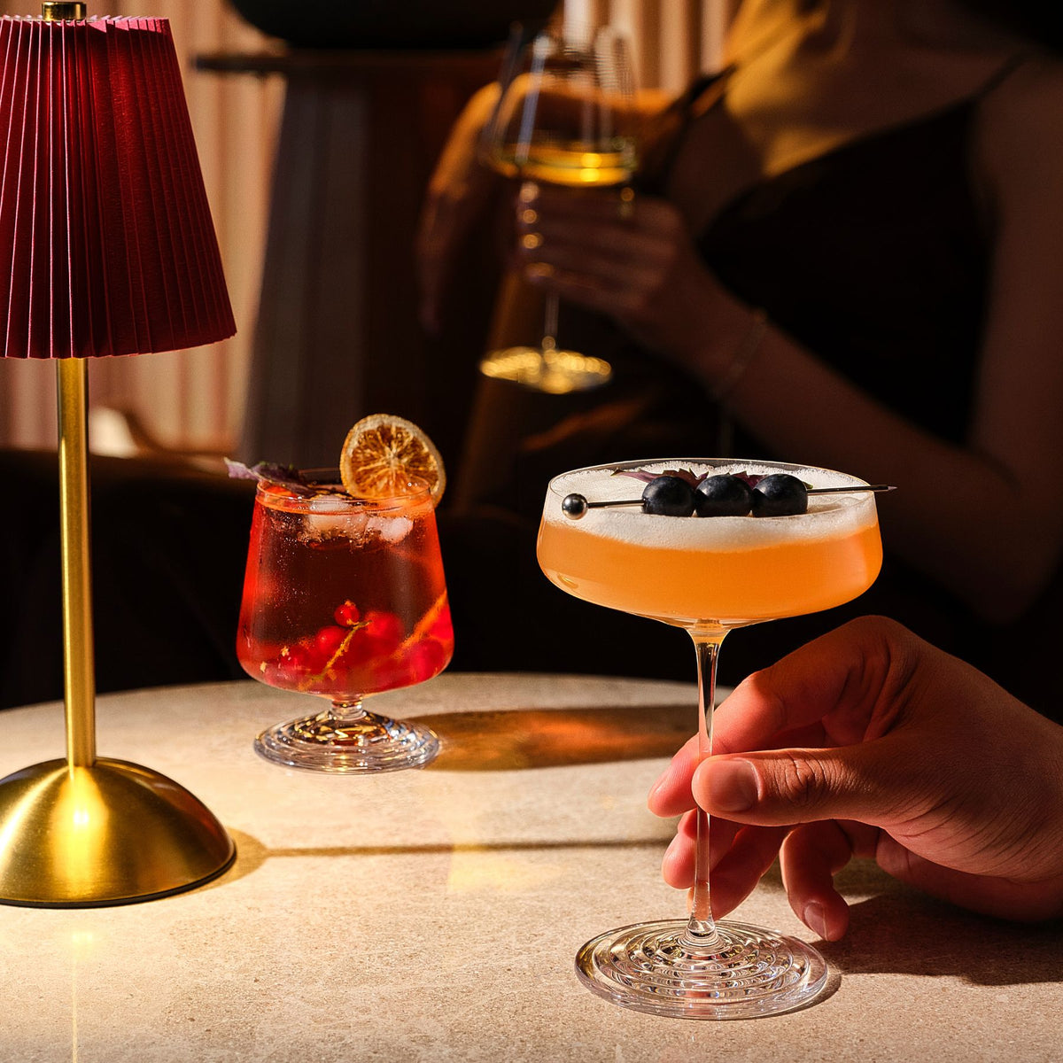 NUDE | Tom Dixon Splash glasses in a dim bar setting: a lowball with a red drink, citrus garnish, and redcurrants beside a red-shaded lamp, and a coupe held by hand with an orange cocktail topped with foam and blackberry garnish.