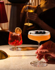 NUDE | Tom Dixon Splash glasses in a dim bar setting: a lowball with a red drink, citrus garnish, and redcurrants beside a red-shaded lamp, and a coupe held by hand with an orange cocktail topped with foam and blackberry garnish.